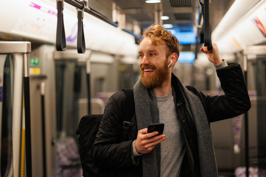 Happy male subway passenger is standing in the train wearing wireless headphones and a smartphone in his hand